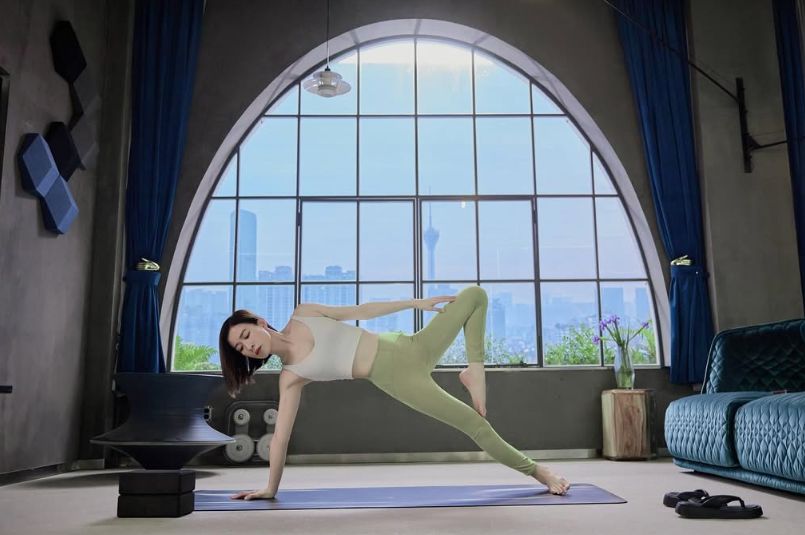 身穿白色運動背心及淺綠色瑜伽褲的女士在落地大窗前練習側平板，展現緊緻身材與凍齡體態。
